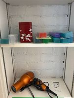 Shelf with various medication holders, one red floral canister and one other can, with an orange Conair hair dryer below.