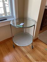 Round glass top side table with frosted glass bottom shelf and chrome metal frame. Photo taken from standing angle showing entire table and wheels on hardwood floor.