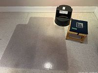 Black plastic step stool, wicker stool with cushion, and transparent plastic carpet protector laid out on floor