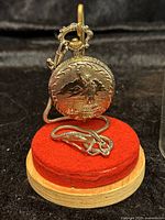Front view of gold-tone quartz pocket watch featuring Statue of Liberty design resting on red velvet stand with matching chain coiled beside it.