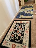 View of two area rugs on floor, black floral rug in foreground, blue floral rug in background