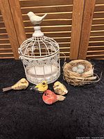 White decorative bird cage with intricate design and white bird on top, four painted bird figurines in yellow, red, tan, and brown colors, natural twig bird nest holding three small stone eggs.