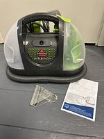Bissell Little Green portable cleaner model 3369 seen from the front, showing black and green body with built-in hose and handle. Attached power cord and a transparent cleaning attachment placed beside it, along with the owner's manual.