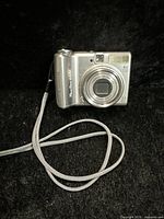 Front view of Canon PowerShot A570IS digital camera showing lens, flash, viewfinder, and model branding with attached wrist strap on black background.