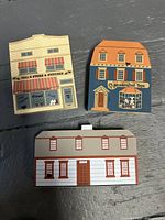 Three Cats Meow Village collectible wooden buildings arranged on a black surface showing front facades: Nell's Stems & Stitches, Royal Langford Jones, Abigail Adams Birthplace.