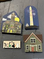 Four wooden Cats Meow Village collectibles arranged on a dark wood surface: Spooky Ridge Courthouse, Washington Monument, Trick or Treaters children, and Vandenberg House. Each painted with night theme, and includes a hidden black cat figure.