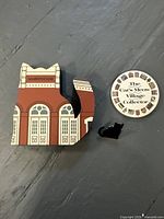 Wooden Markethouse building piece, circular collector's pin with logo, and black cat pin displayed on a grey surface