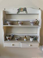Photo of white wooden display shelf decorated with small floral painting, holding seven miniature teacups and vessels each with colorful parrot figurines on or near the rims.