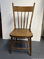 Front and angled front views of a wooden farmhouse spindle back chair showing carved design on backrest, turned spindles, contoured wooden seat, and turned legs and stretchers.