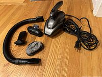 Full view of the vacuum cleaner with all accessories arranged on a wooden floor: the main vacuum unit, flexible hose, flat nozzle, small brush attachment, and filtering bag.