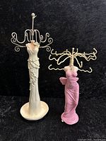 Two figural women jewelry display stands, white and pink, with wire hooks to hang jewelry, against a black background.