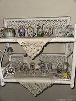 White wicker shelf with crocheted lace doilies on two shelves, displaying an assortment of miniature porcelain vases and decorative ceramic pieces, all featuring floral designs and pastel colors.