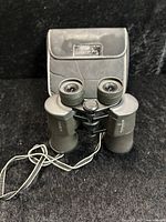 Front angled view of Minolta binoculars with neck strap and carrying case in background on black surface.