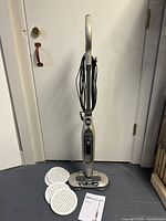 Full view of upright steam mop standing against wall with three round pads and owner's guide laid on floor.