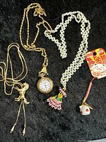 Full view of four jewelry items on black background: beaded flower necklace, horse pendant necklace, pocket watch pendant necklace, and non-metallic charm on cord.