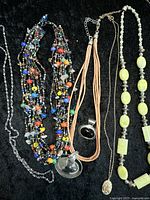 Full view of the lot showing colorful multi-strand necklace, tan cord pendant necklace, silver-tone chain necklace, and pale yellow bead necklace on a black background.