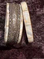 Two bangle bracelets side by side on dark textured surface, one mother of pearl inlaid and one metal with embossed pattern.
