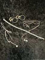 Full view of assorted jewelry pieces including clip hoop earrings, clear stone bracelet, and delicate chain necklace with small clear stone, displayed on black surface