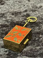 View of rectangular brass pillbox with orange enamel top featuring umbrella and raindrop designs, attached chain and keychain fastener shown lying on dark fabric.