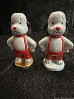 Two ceramic dog figurines of character Sweep, one with a gold base and one with white base, standing side by side on black background