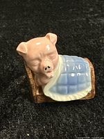 Front angle view of Priscilla Pig figurine sleeping in a bed with blue blanket