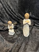 Two Willow Tree angel figurines on black fabric background. Smaller figurine kneeling, larger standing, both have wire wings.