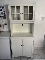 Full front view of the white kitchen pantry cabinet showing the closed glass-paneled upper doors, open middle shelf area, and closed bottom solid doors.
