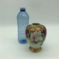 The vase standing next to a water bottle to show scale, bulbous shape, white and pink hand painted roses with green leaves, gold accents on top and bottom, gold beading detail, felted bottom.