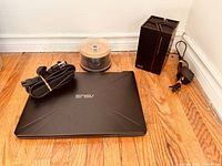 Laptop closed with power cables, wireless repeater, and CD spindle on wood floor