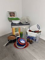 Wide shot showing all cooking and camping items arranged on floor against wall including stacked plates, bowls, mugs, glasses, cookware, utensils, storage containers and tablecloth.