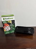 Photo of FoodSaver vacuum sealer machine next to a box of vacuum seal rolls, showing the front and control buttons of the sealer and packaging details of the seal rolls.