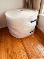 Large commercial roll of bubble wrap standing upright on a wooden floor near a curtain.