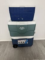 Three coolers stacked on each other against a white wall on wooden floor: Blue Super Cooler on top, green Igloo Island Breeze in middle, and blue Coleman cooler with wheels at the bottom.