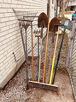 All garden tools arranged upright against a building wall, showing rakes, shovels, and a broom