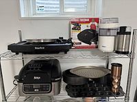 Wide view of wire rack shelf with Starfrit indoor grill on top with pizza pan and metal travel mug, Ninja air fryer on second shelf with baking pans and mug