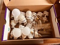 Top view of open box showing mix of round LED bulbs and spiral CFL bulbs with standard screw bases, some clear glass candle-style bulbs visible.