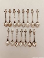 Photo displaying the entire lot of 14 vintage collector spoons arranged in two rows, each with a detailed bust of a Canadian Prime Minister on the handle and engraved years in the bowl.