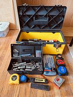 Open large yellow Stanley toolbox showing the smaller black toolbox and some tools inside, along with various tape measures, drill bit sets, and hand tools arranged outside in front of the box.