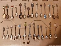 Full lot of 29 vintage collector spoons laid out in two rows with a ring and pin among them.
