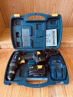 Blue hard plastic carrying case open showing cordless drill, two batteries, charger, and instruction manual