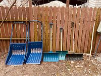 Snow shovels and snow scoops lined up against a wooden fence