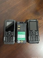 Two Sonim two-way radios and one battery pack on a wooden surface.