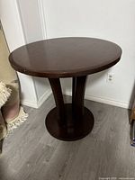 Front angle showing round wooden table with three-support pedestal base and circular base, on light wood floor near white walls and fabric items.