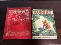 Photo of two vintage children's books on table: 'The Sleeping Beauty' with red decorative cover and 'Rupert The Daily Express Annual' with illustrated bear cover.