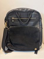 Front view of the black leather backpack showing the front suede panel and zippered pockets with shoulder strap relaxed to side.