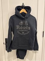 Front view of the black Roots hoodie and sweatpants hanging together, showing logo on hoodie