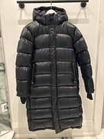 Full front view of the long black TNA down-filled puffer coat with hood hanging on a hanger