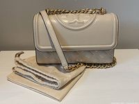 Front view of cream leather bag with quilted lower exterior and logo-embossed flap, dust bag folded beside it