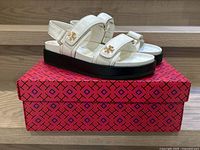 Side view of Tory Burch Kira Sport sandals in ivory goat leather with black platform soles, showing adjustable straps and gold-tone logo accent. Sandals placed on red patterned box.