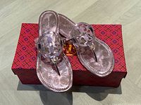 Pair of Tory Burch pink mirrored metallic sandals on top of their red patterned box.